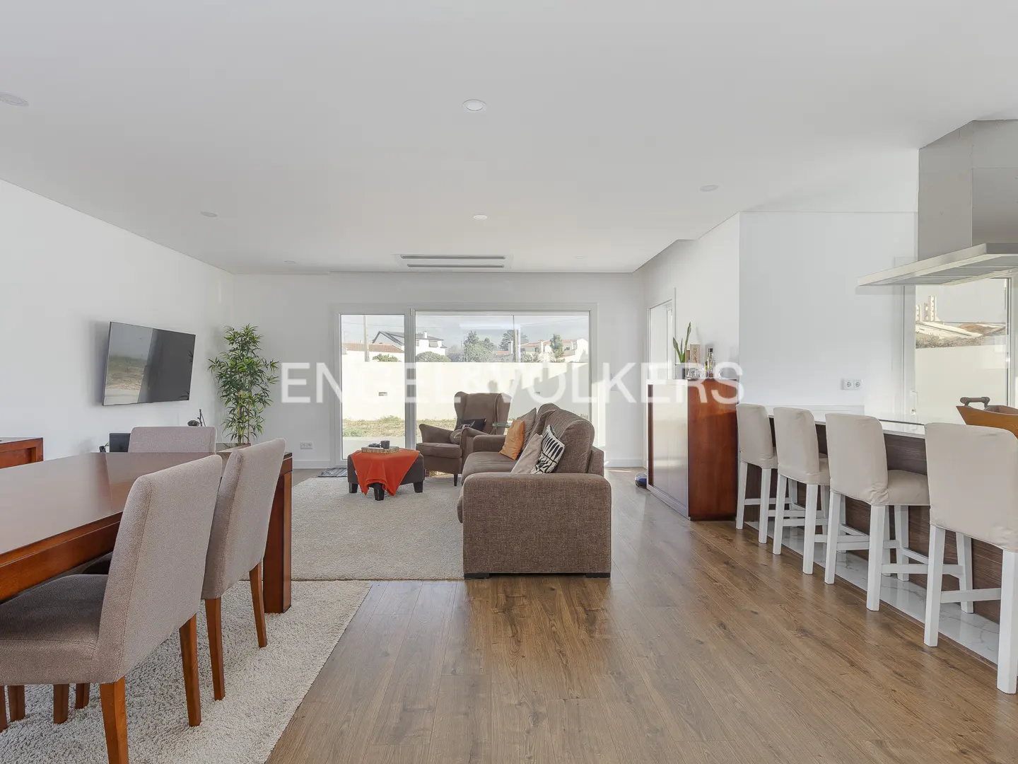 Open-concept living space with wood floors, white walls, and a dining table with beige chairs. A brown sofa and armchair sit on a cream rug near a large window.