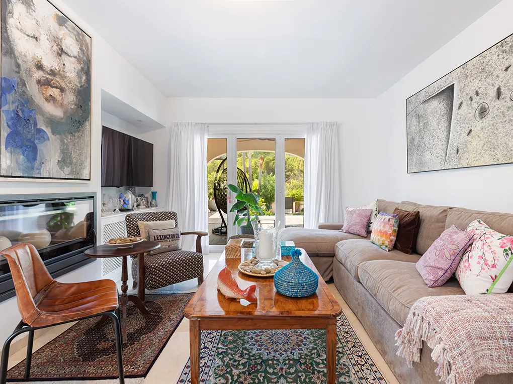 Living room with a brown sectional sofa, wooden coffee table, and patterned rug. Artwork hangs on the white walls. Patio doors lead to a green outdoor space.