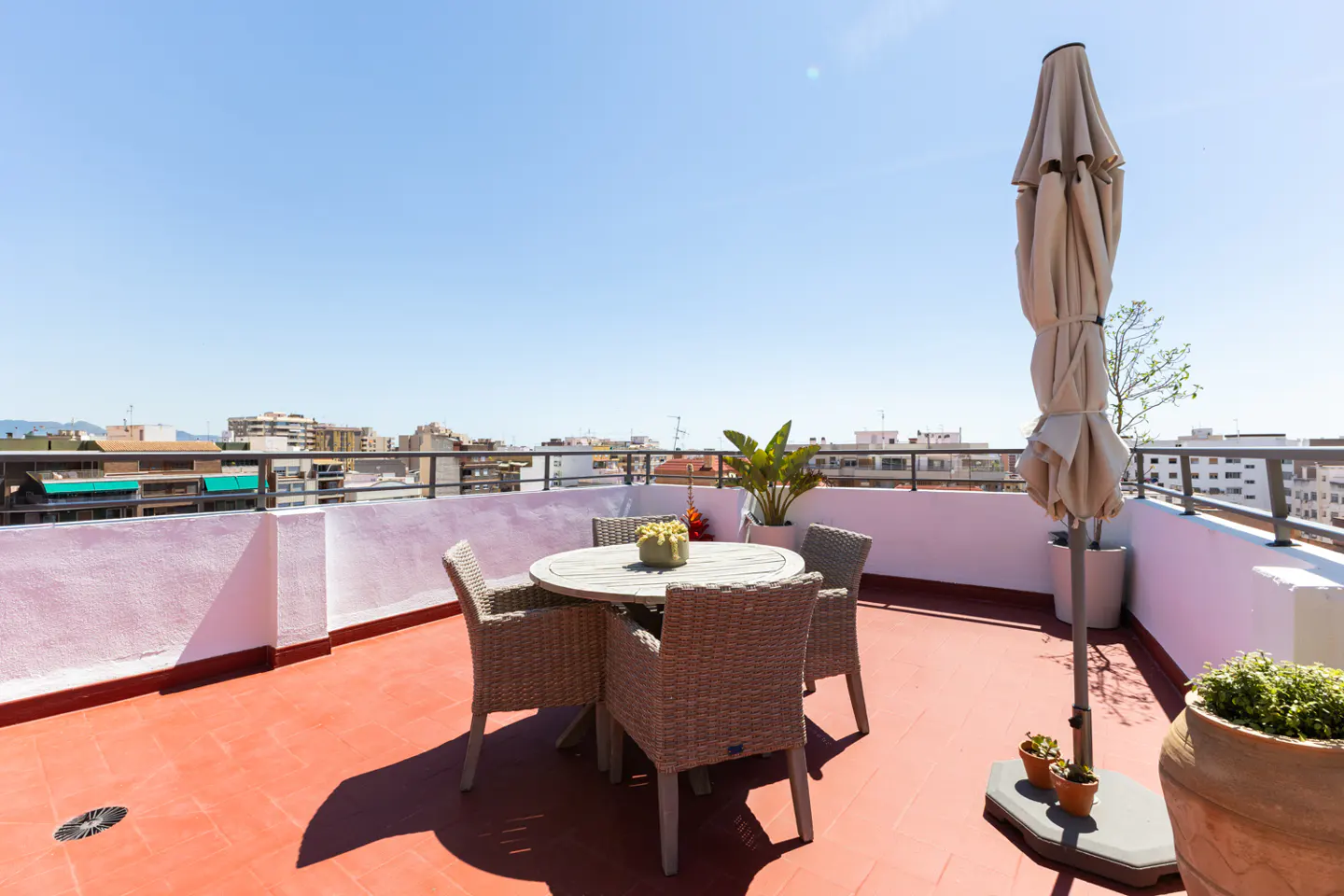Rooftop patio with a round table, four wicker chairs, and a closed beige umbrella. City buildings are visible in the background under a clear blue sky.