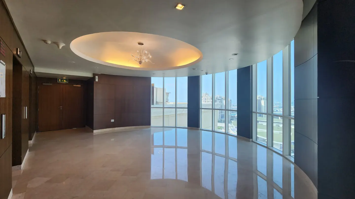 Bright, empty room with marble floors and curved floor-to-ceiling windows showing a city view. A chandelier hangs from a recessed ceiling.