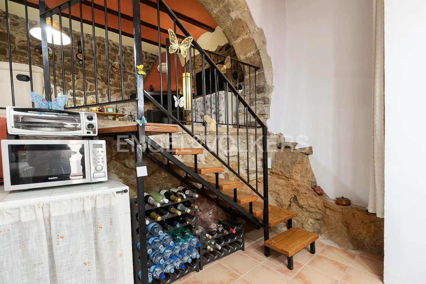 Interior view of a kitchen with a staircase, wine rack, microwave, and toaster oven. The staircase has black metal railings and wooden steps. Butterflies decorate the railings.