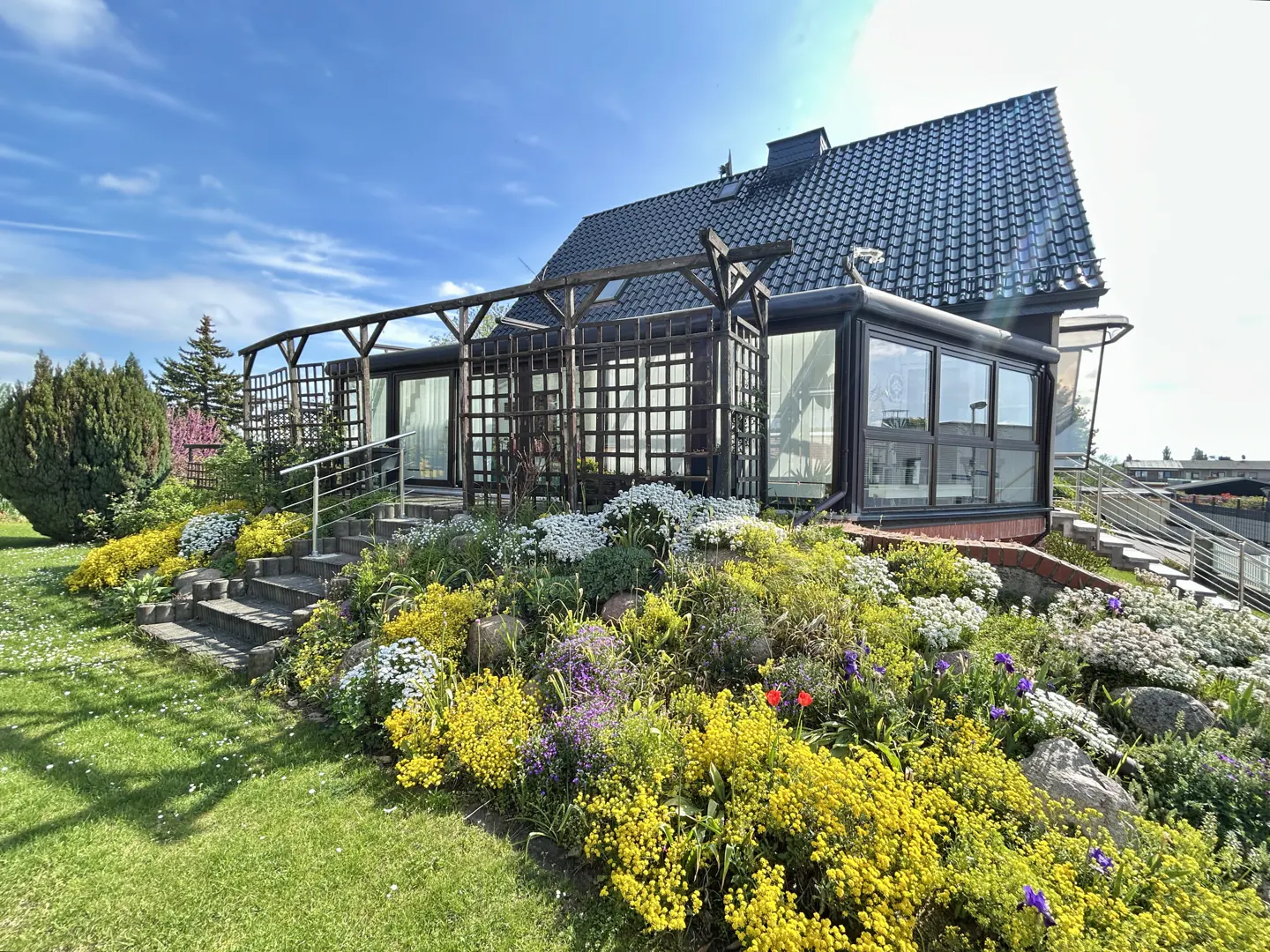 Exterior view of a house with a black roof, a sunroom, and a garden with yellow, white, and purple flowers.