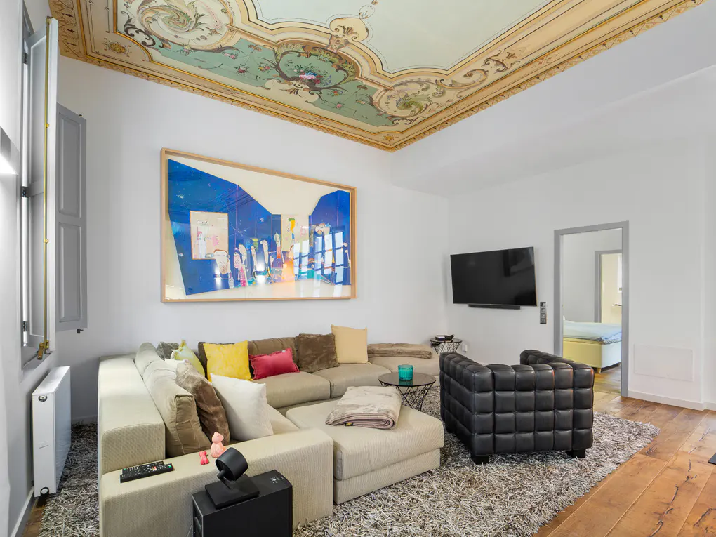 Living room with ornate ceiling, large blue art, and sectional sofa. A black leather chair sits on a gray rug. TV on the wall.