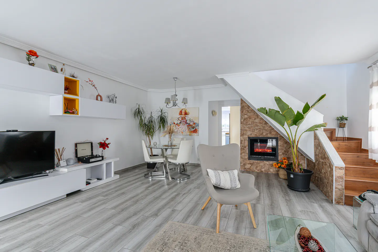 Bright living room with gray wood floors, white walls, and a stone fireplace. A gray armchair sits near a glass coffee table. Dining area and stairs are visible.