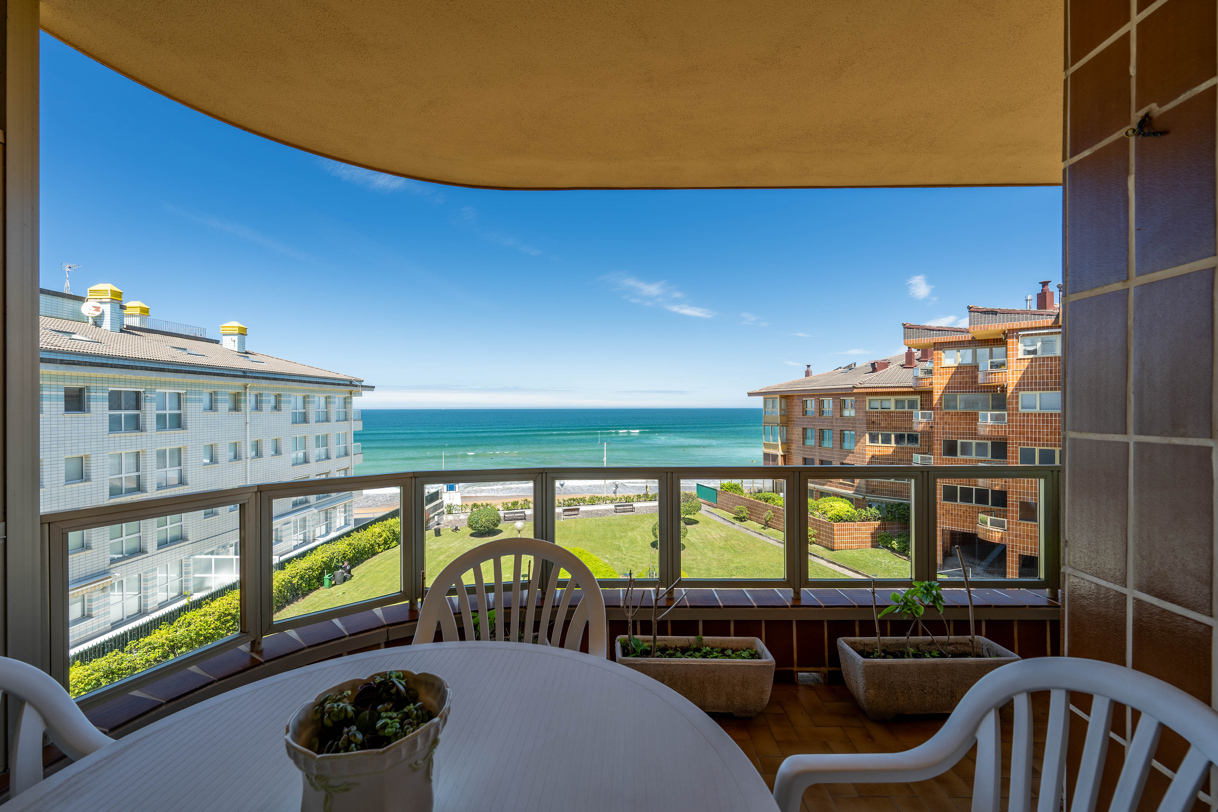 Balcony view with a white table and chairs, overlooking a green lawn, nearby apartment buildings, and the blue ocean.