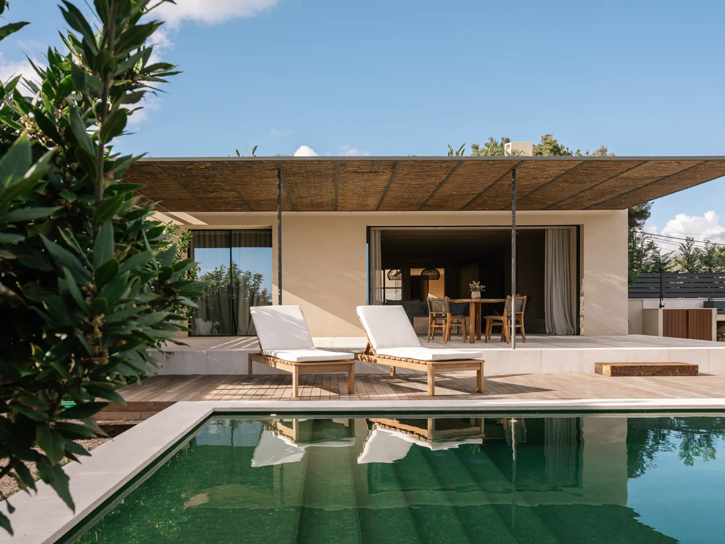 Modern home exterior with a green pool, wooden deck, and two white lounge chairs under a woven shade. Blue sky above.