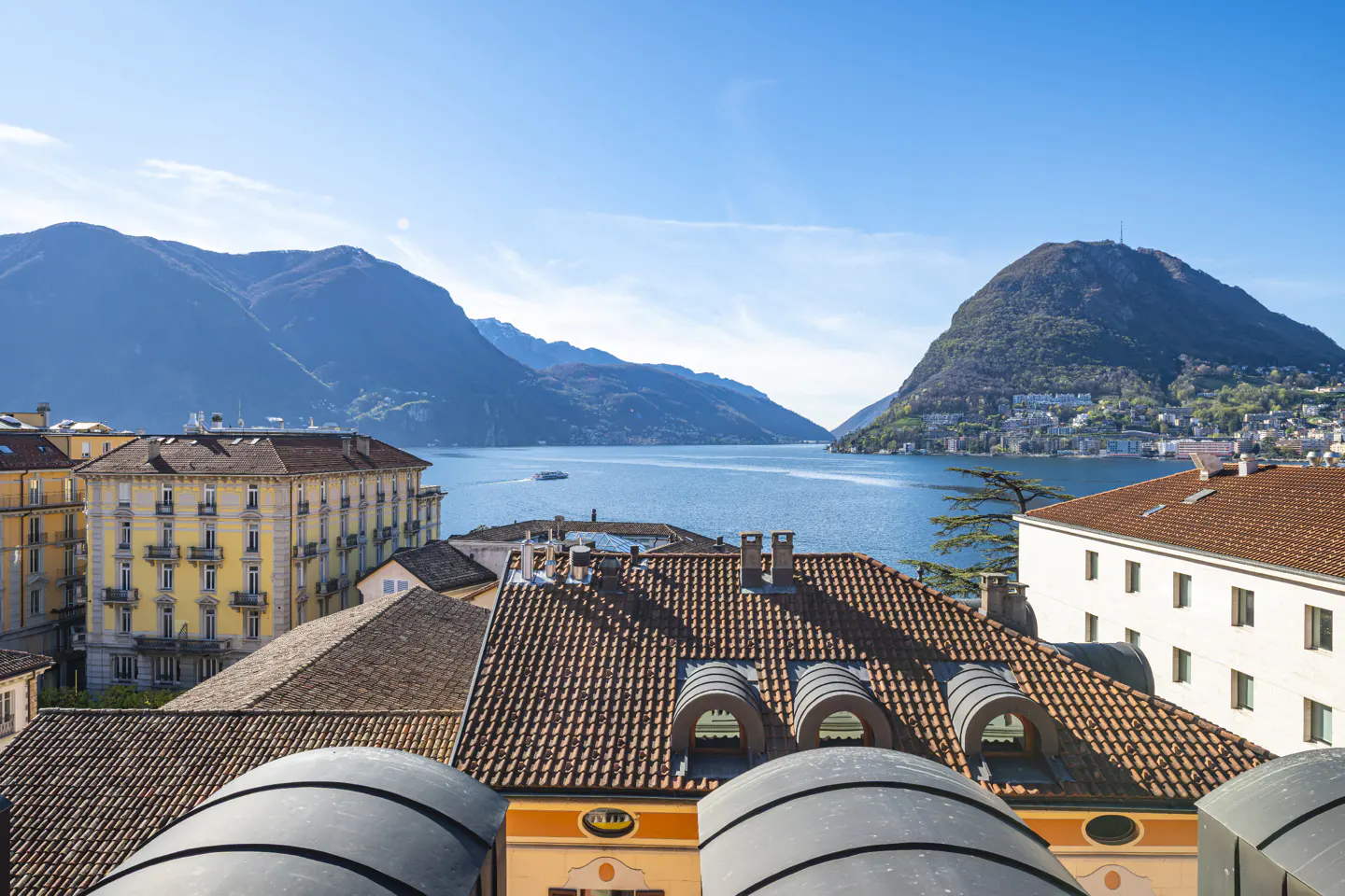 View of Lugano lake and mountains from rooftops with terracotta tiles and yellow buildings under a clear blue sky.