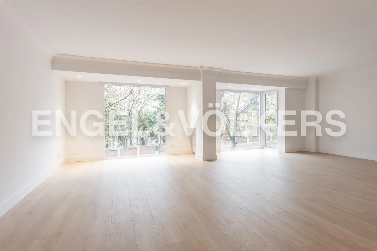 Bright, empty room with light wood floors, white walls, and two large windows overlooking trees. Real estate company logo overlayed.