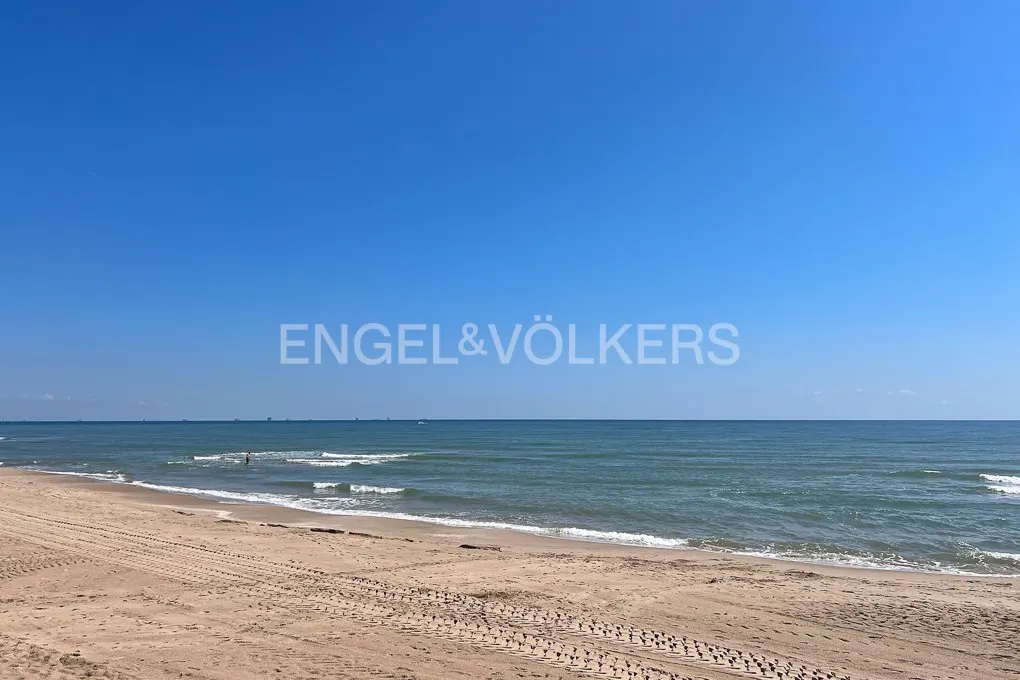 Beach scene with tan sand, blue-green ocean, and clear blue sky. Tire tracks mark the sand. Engel & Völkers logo is superimposed.