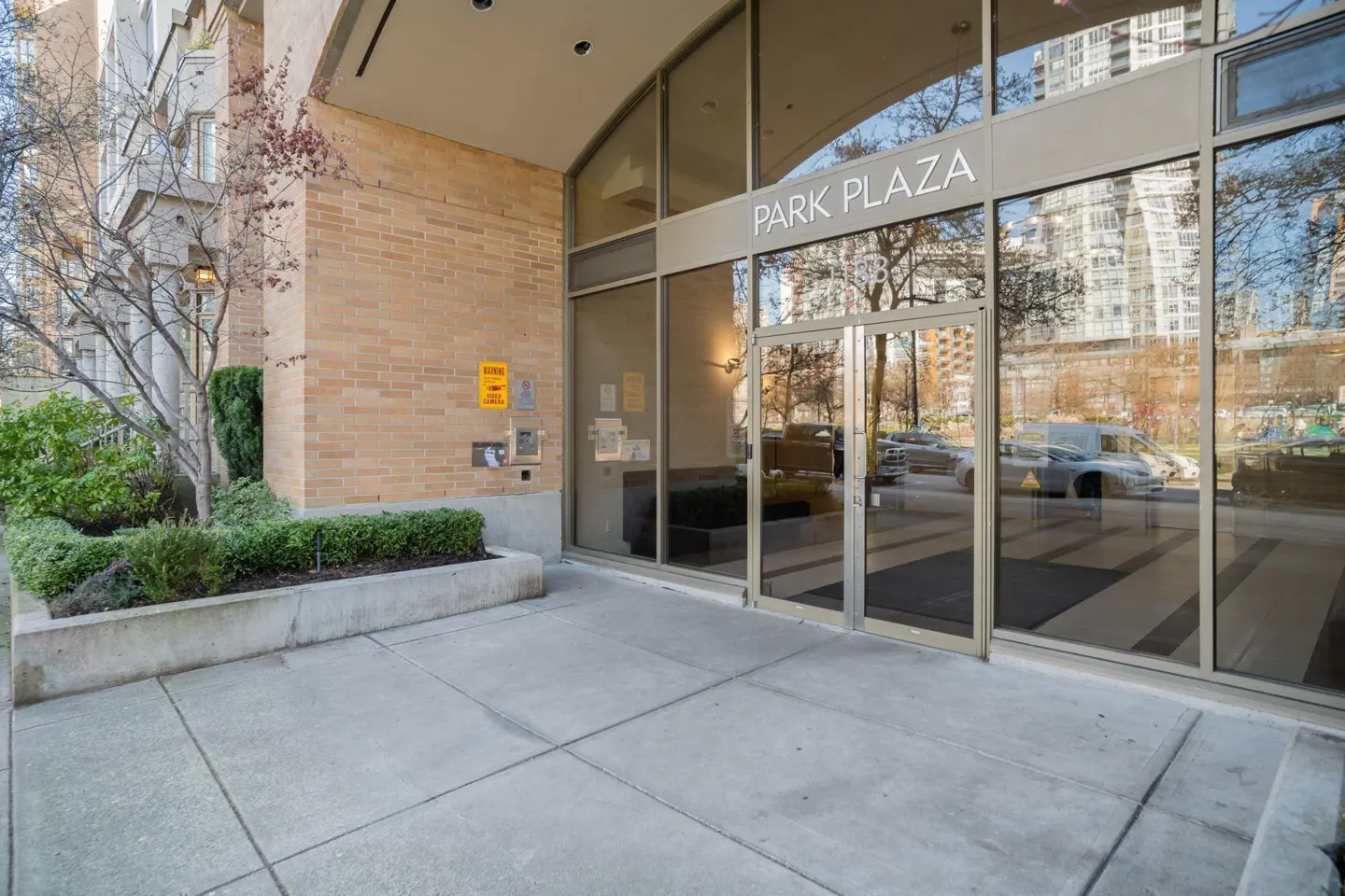 Exterior view of Park Plaza building entrance with glass doors and brick facade. Bushes and trees are planted in front.