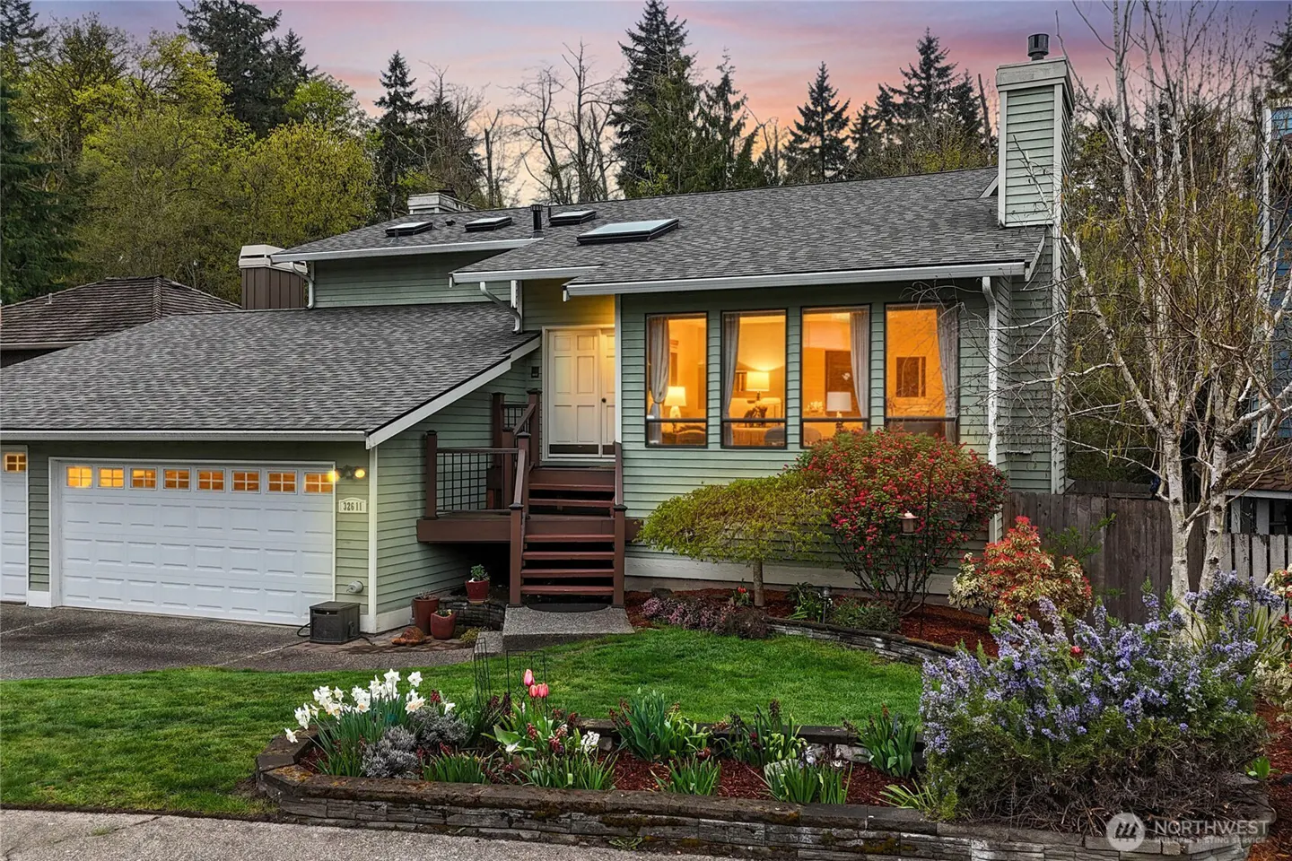Exterior of a two-story, light green house with a gray roof, white garage door, and a lawn with flowers.