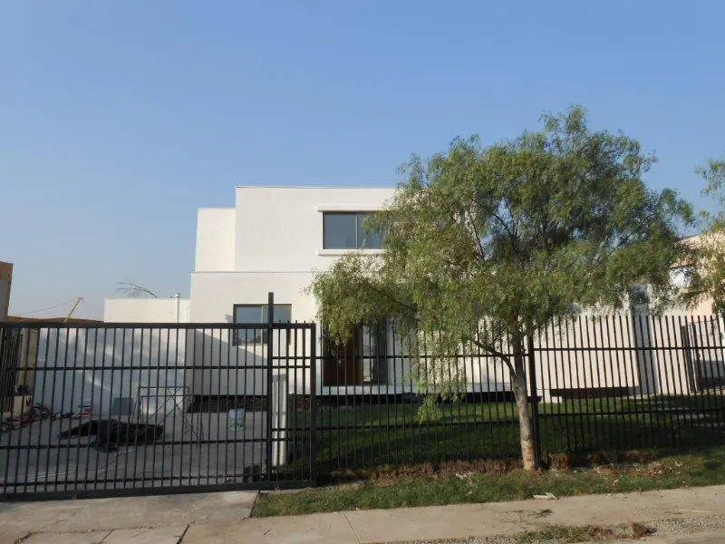 A modern, two-story white house is behind a black metal fence and a green tree under a blue sky.