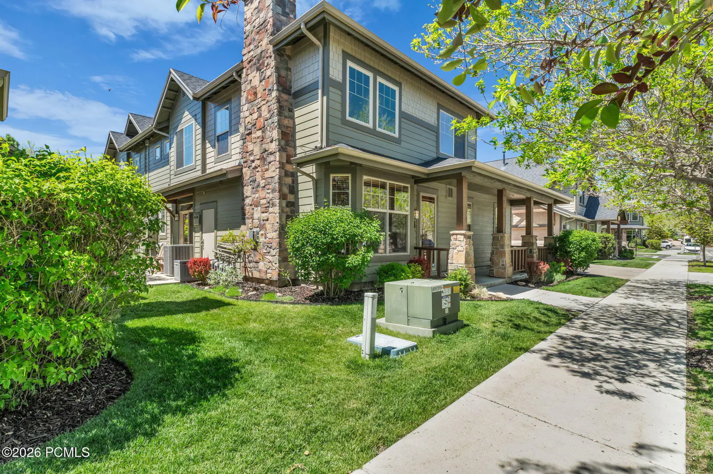 Two-story townhouse with olive green siding, stone chimney, and a front porch, set on a lawn beside a concrete sidewalk.