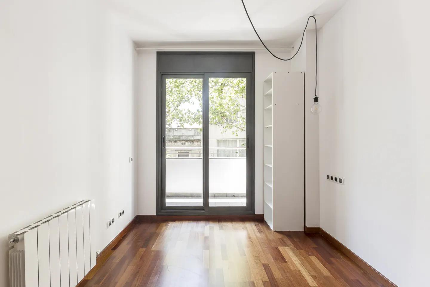 Bright, empty room with hardwood floors, white walls, and a sliding glass door to a balcony. A white bookshelf and a hanging lightbulb are visible.