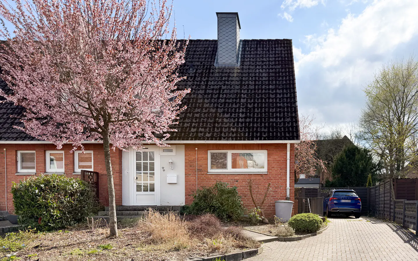 A red brick house with a dark roof and a blooming pink tree in the front yard on a sunny day.
