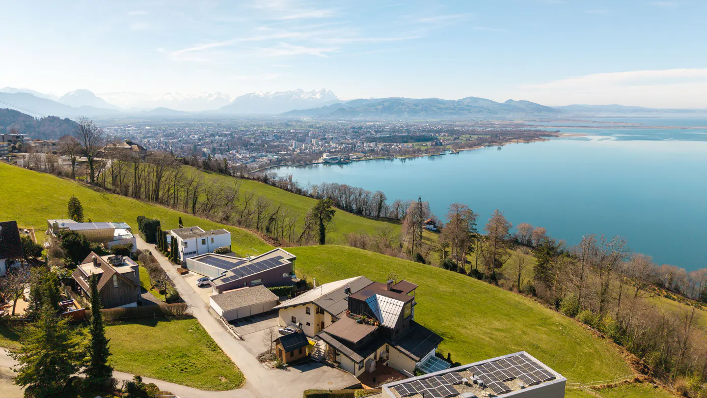 Aerial view of houses on a green hillside overlooking a blue lake, a city, and snow-capped mountains under a bright sky.