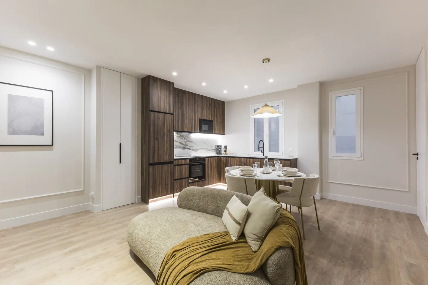 Modern open-plan living area with a curved beige sofa, dining table, and dark wood kitchen cabinets on light wood floors.