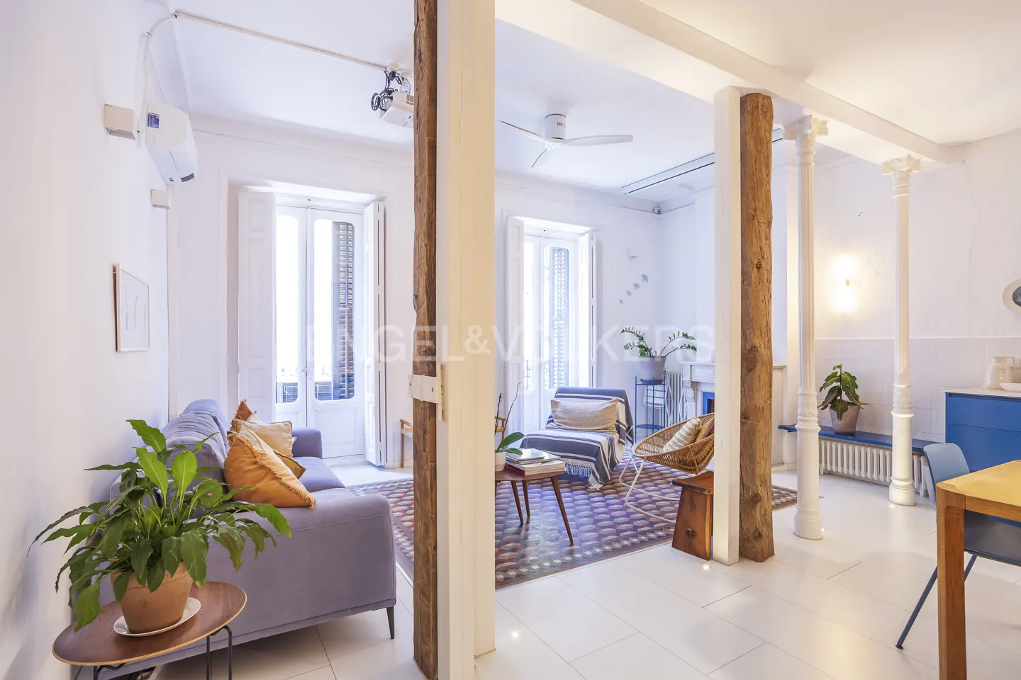 Bright living room with a blue sofa, wooden beams, and white walls. Plants add a touch of nature to the space.