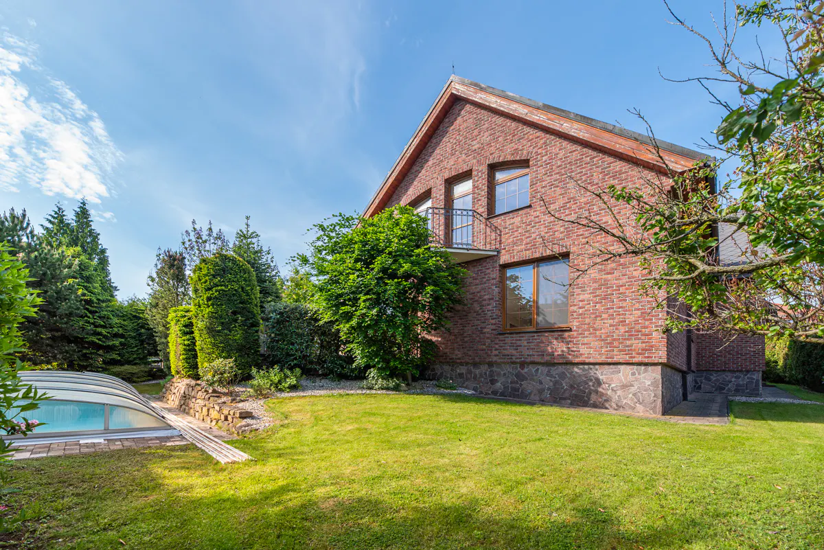 Brick house with a balcony, green lawn, and a covered pool on a sunny day.
