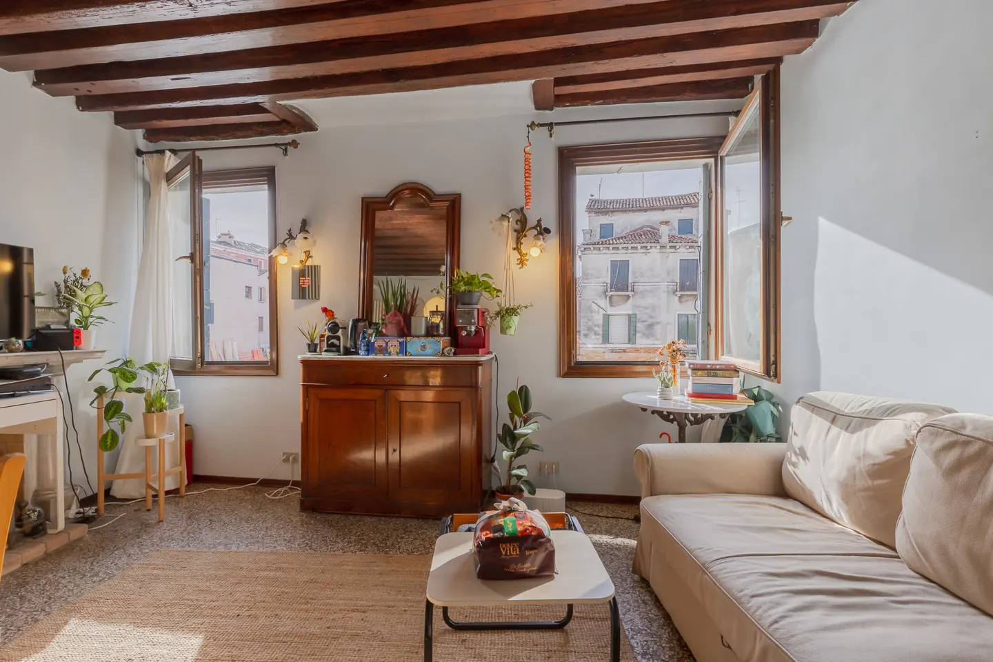 Living room with exposed wood beams, white walls, and two windows. A brown cabinet with a mirror sits between the windows. A beige sofa and coffee table are in the foreground.
