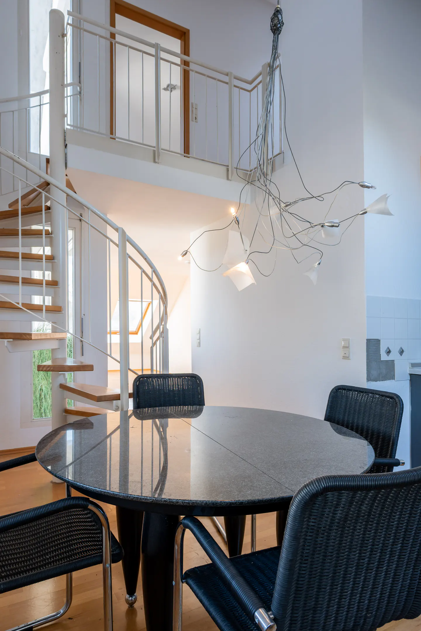 Interior view of a dining area with a round, black table and chairs. A spiral staircase and modern chandelier add architectural interest.