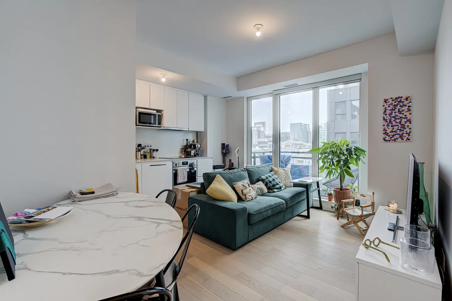 Bright condo interior with a white kitchen, teal sofa, and a marble table. Large windows offer a city view.