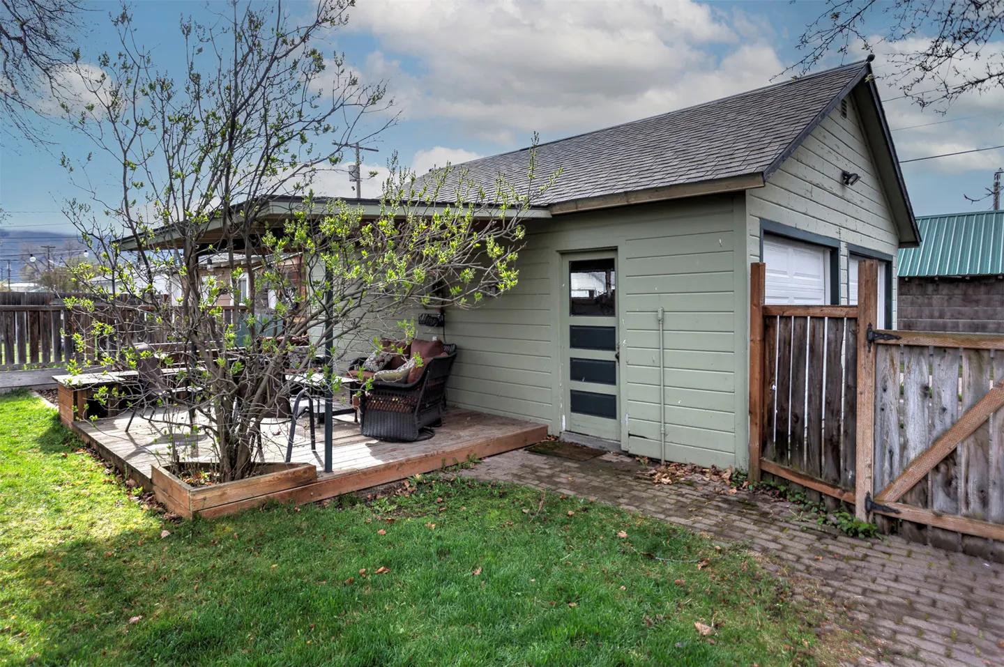Exterior view of a light green house with a deck, chairs, and a tree in the yard. A wooden fence is on the right.