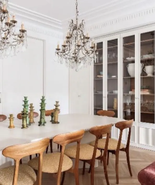 Elegant dining room with a white table, wooden chairs, and glass-front cabinets. Crystal chandeliers hang above. Green and gold decorative objects adorn the table.