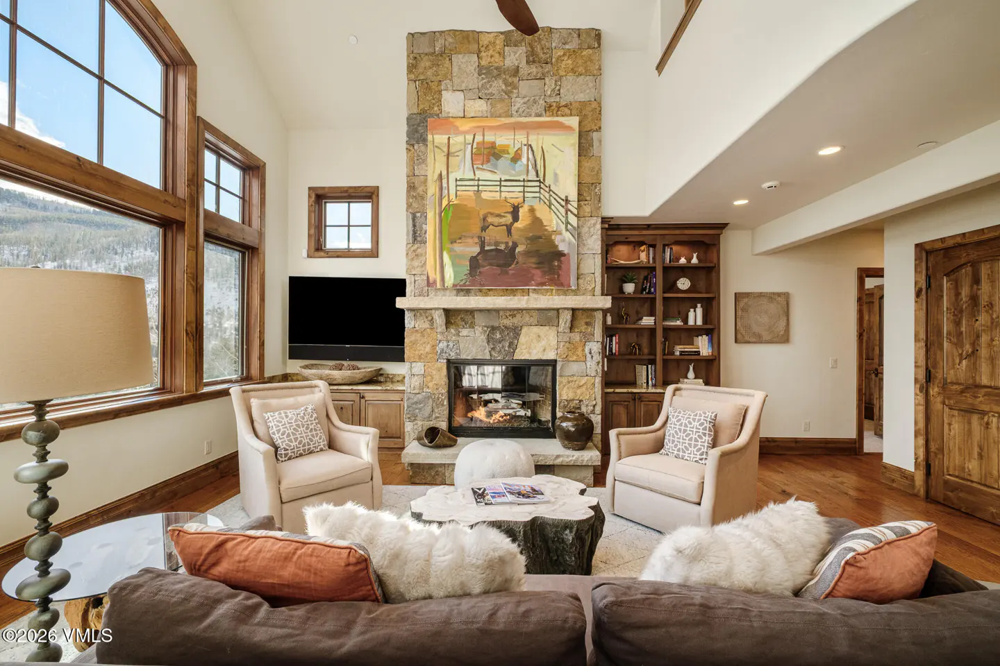 Living room with stone fireplace, abstract art, and large windows. Brown sofa, beige chairs, and a wood coffee table.