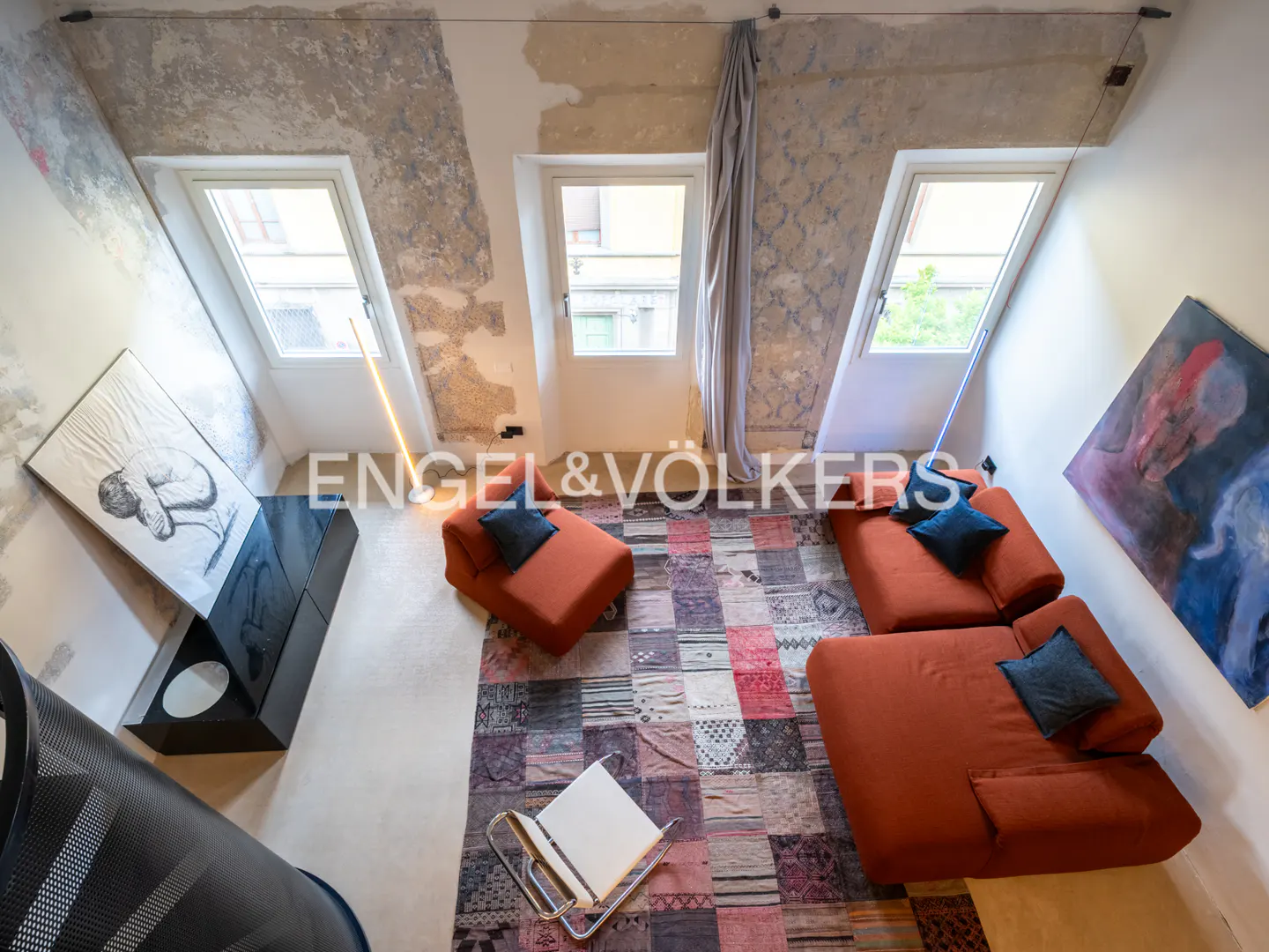 Loft apartment with orange sofas, blue pillows, and a patchwork rug. Three windows let in natural light. Art adorns the walls.