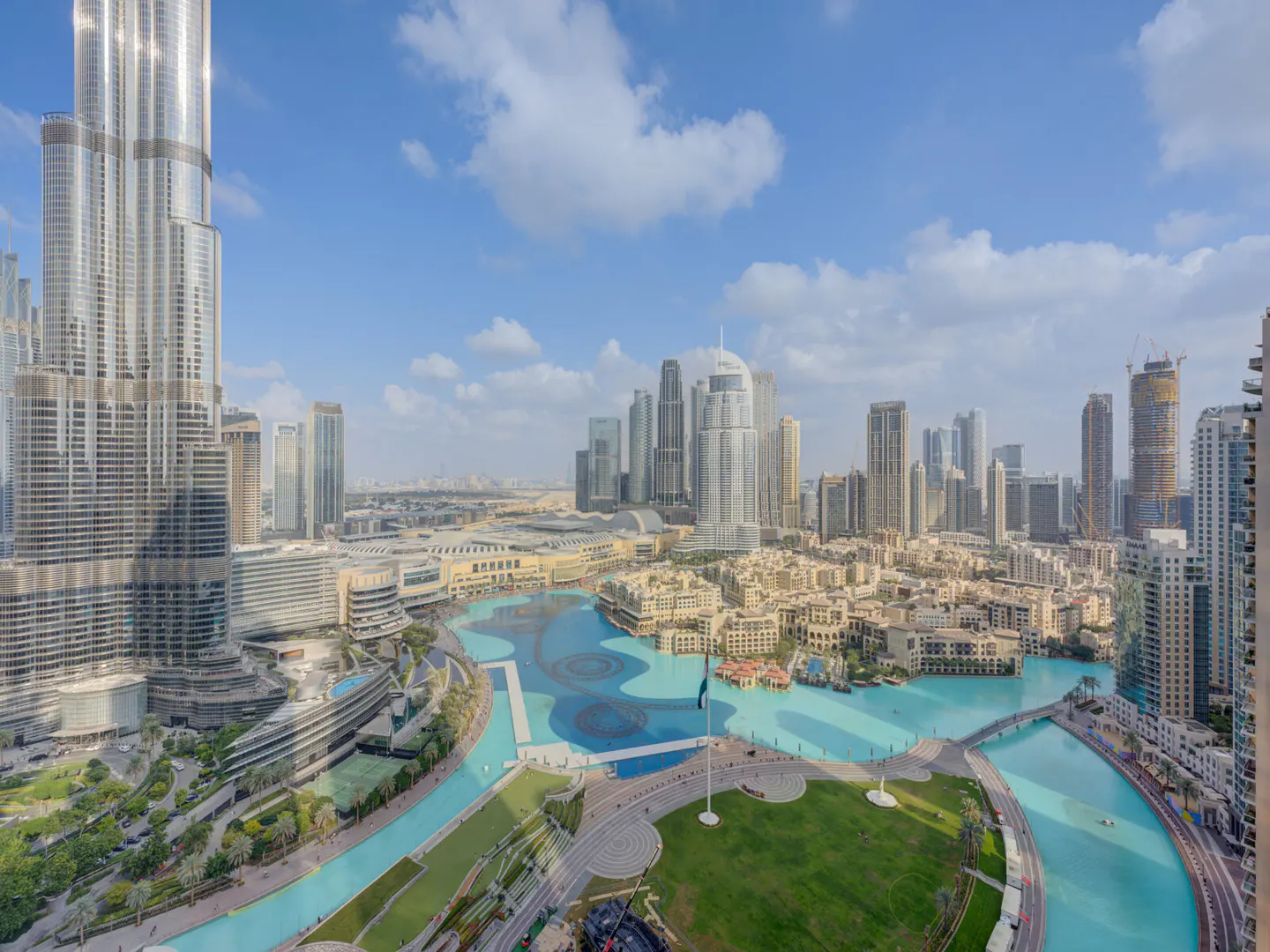 Dubai skyline view featuring the Burj Khalifa, a turquoise canal, green spaces, and other skyscrapers under a blue sky with clouds.