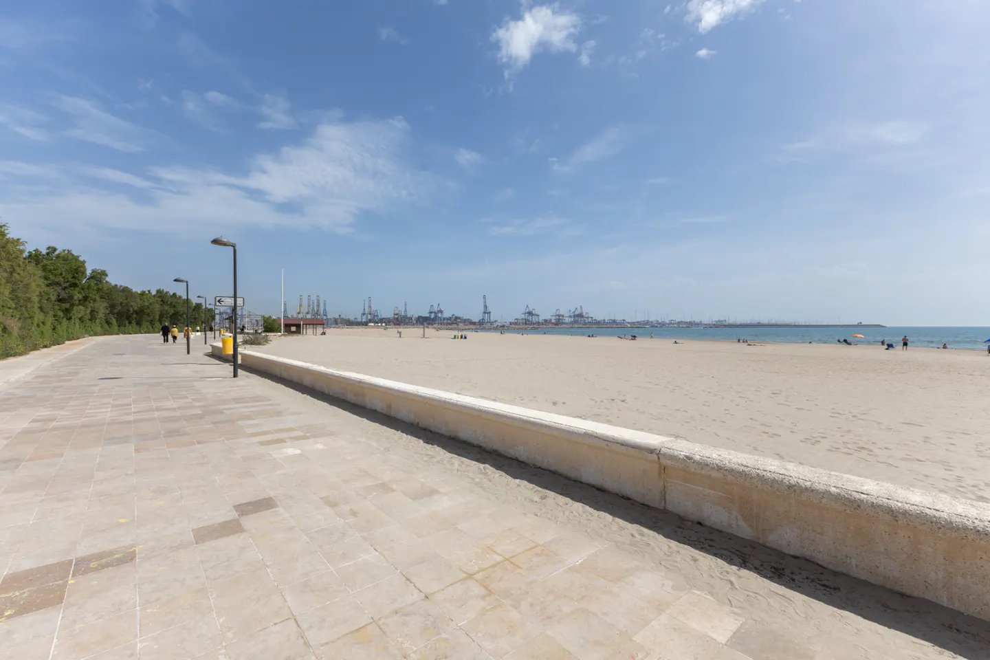 Beach view with a stone walkway, trees, and lampposts on the left. In the background, there is a sandy beach, the ocean, and a port with cranes.