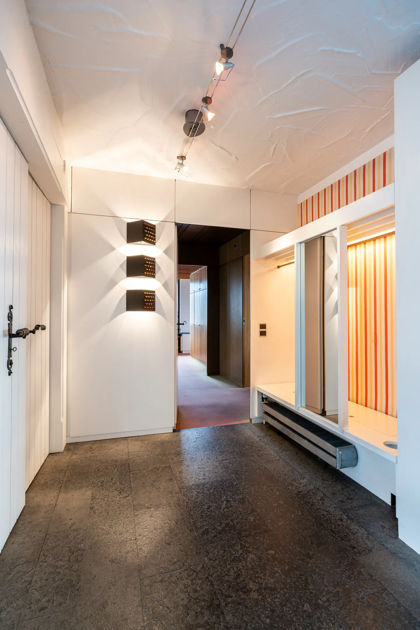 Interior view of a modern hallway with white walls, dark stone floors, and unique lighting fixtures. A doorway leads to a hallway with a red carpet.