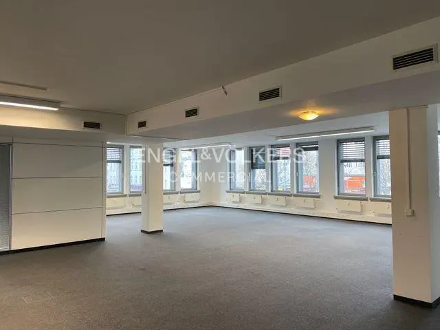 Empty, open-plan commercial office space with grey carpet, white walls, and large windows letting in natural light.