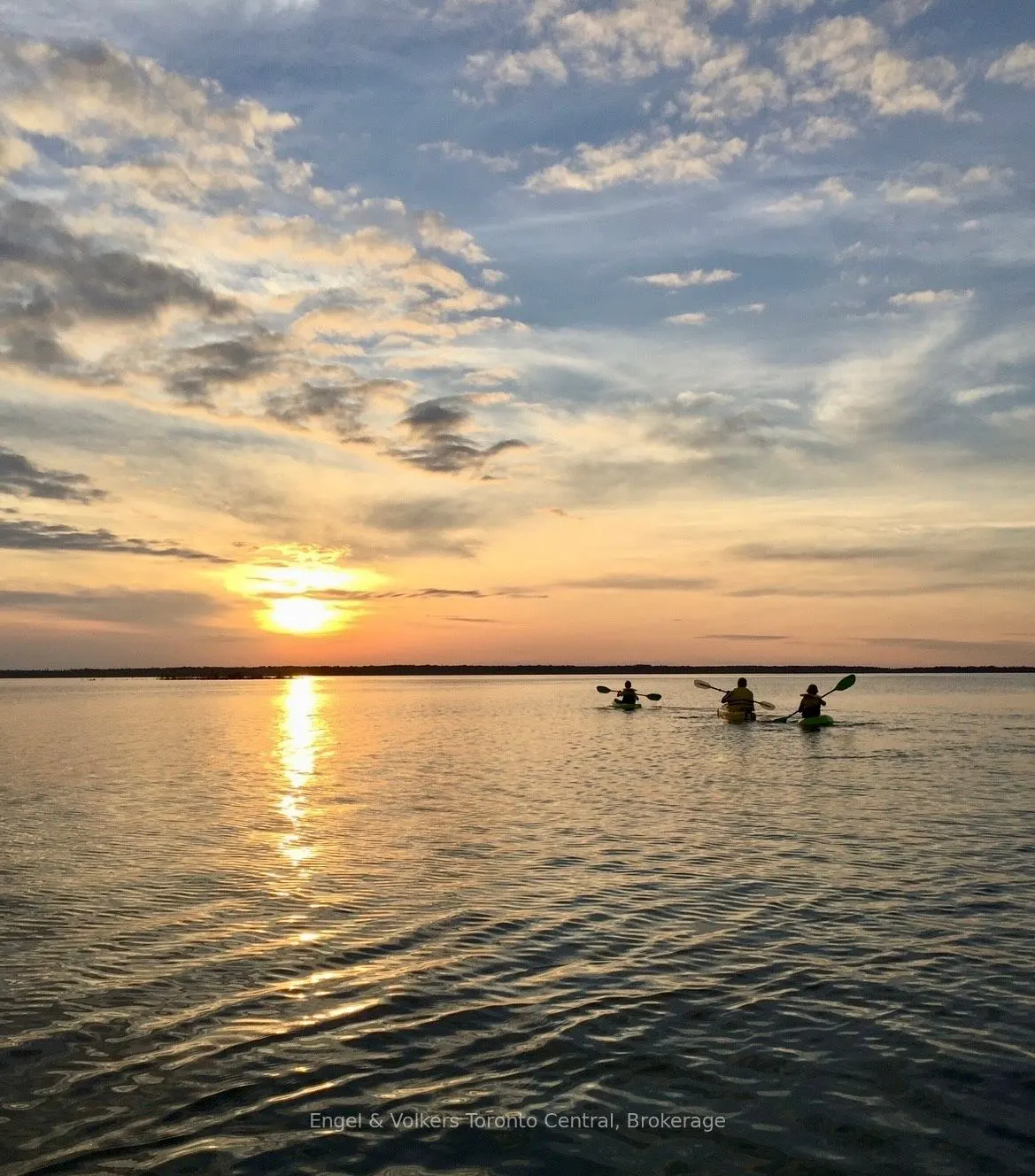 Three people kayak on a calm lake at sunset. The sky is blue and orange with clouds.