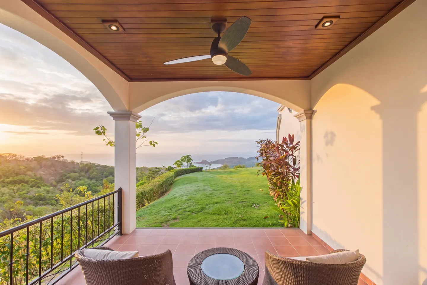 Covered patio with wicker chairs and table overlooks green lawn, trees, and ocean at sunset. Ceiling fan and recessed lights above.