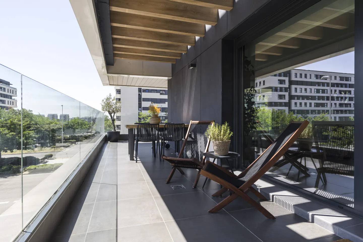 Modern balcony with gray tile flooring, wooden ceiling beams, glass railing, and outdoor furniture overlooking a city.