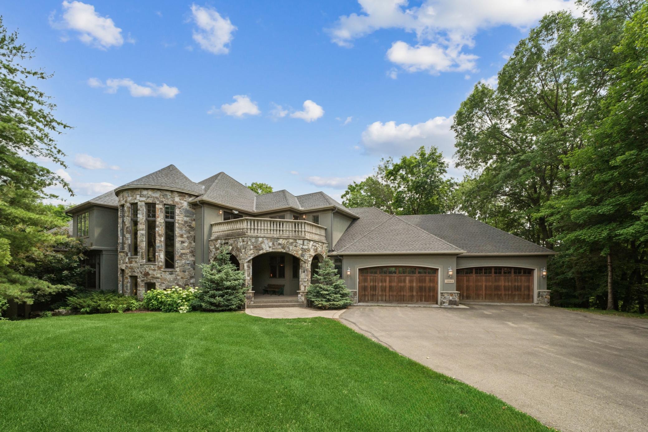 Exterior view of a large, gray house with a stone turret, balcony, and three-car garage, surrounded by green lawn and trees under a blue sky.