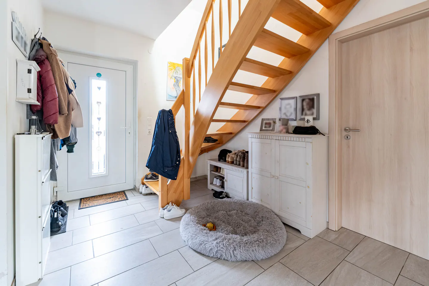 Bright entryway with a white door, wooden stairs, and a gray dog bed on tiled floor. Coats hang near the entrance.