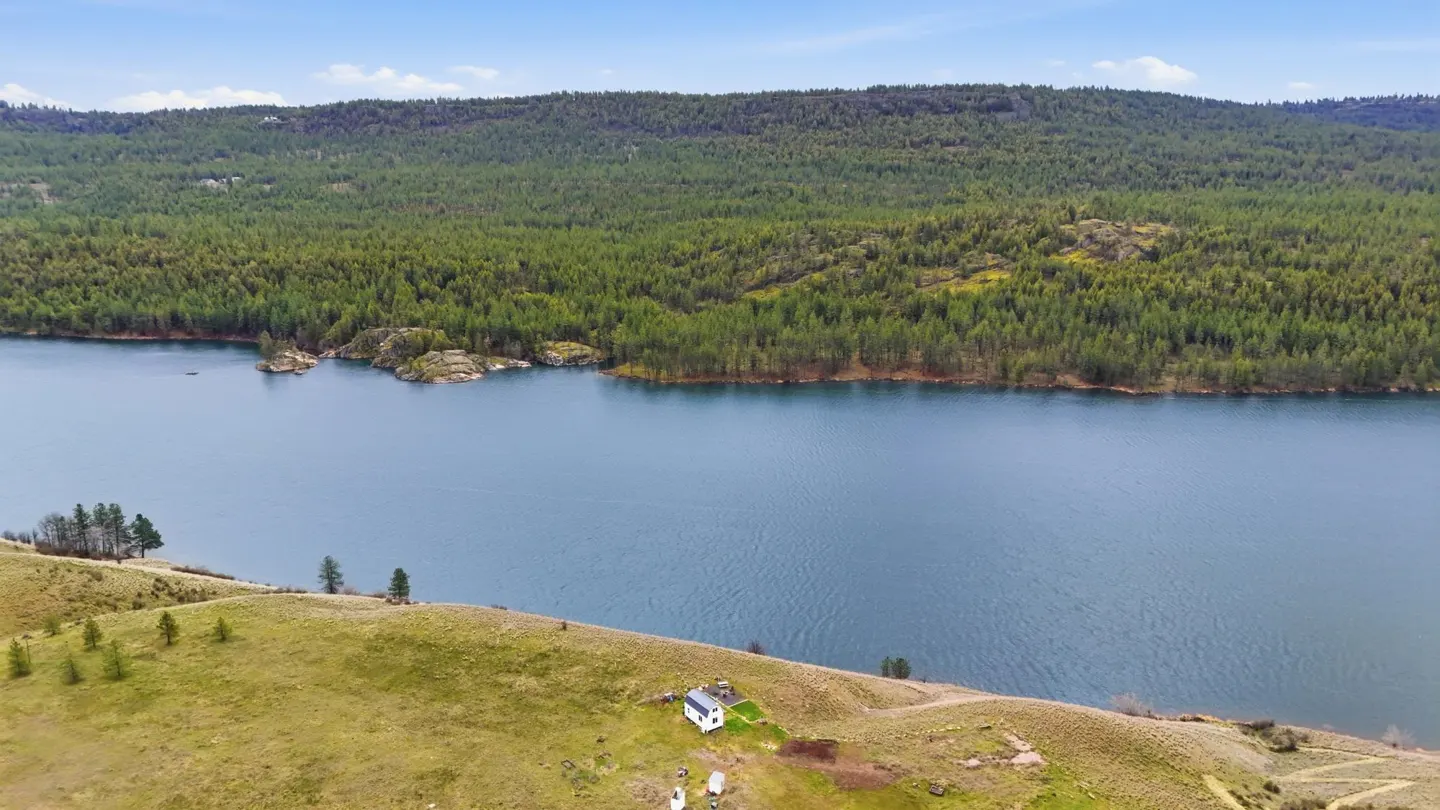 Scenic view of a blue lake bordered by green hills and a dense forest under a blue sky. A small white house sits on the hillside.