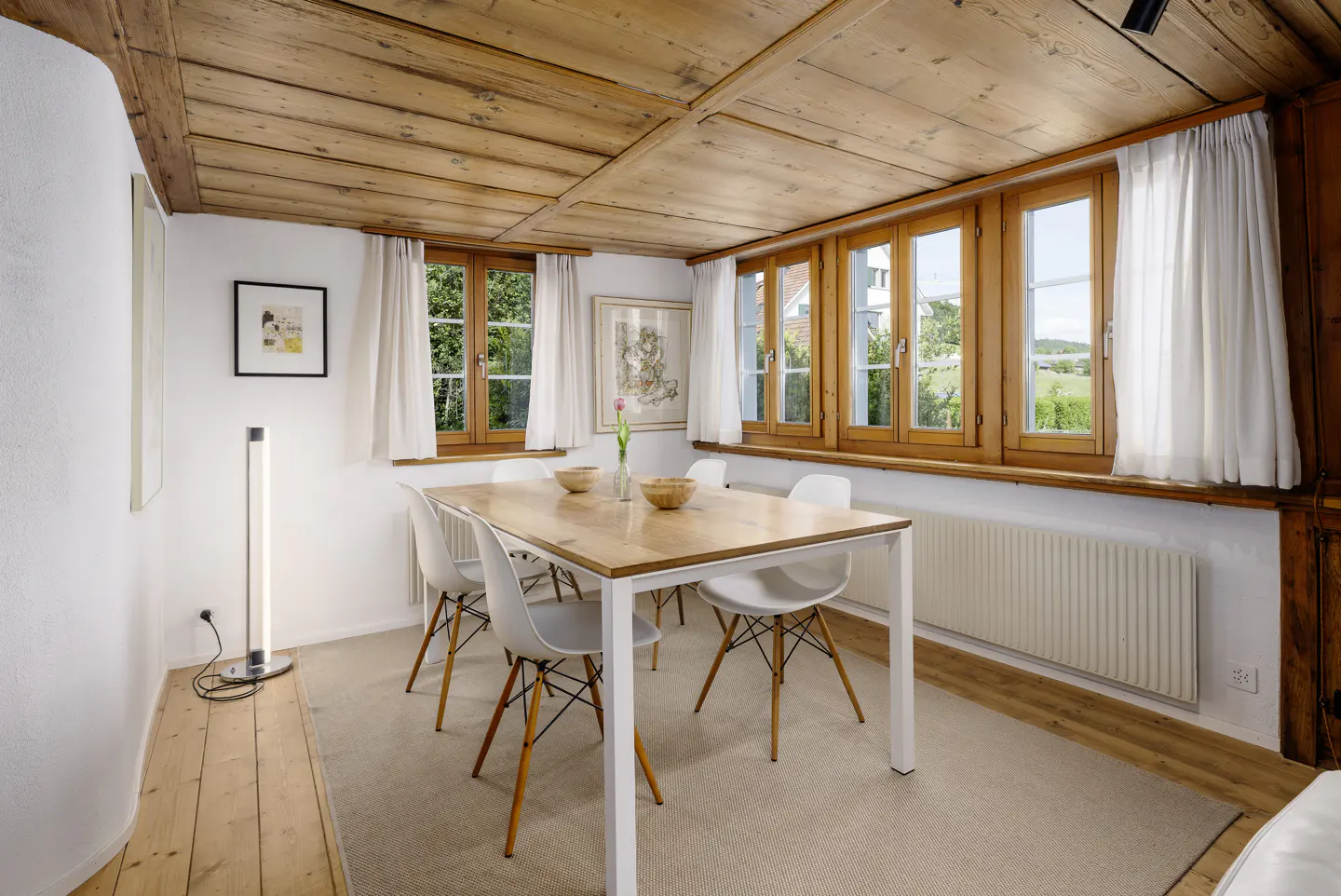 Dining room with wooden ceiling, white walls, a wooden table with four white chairs, and windows overlooking greenery.