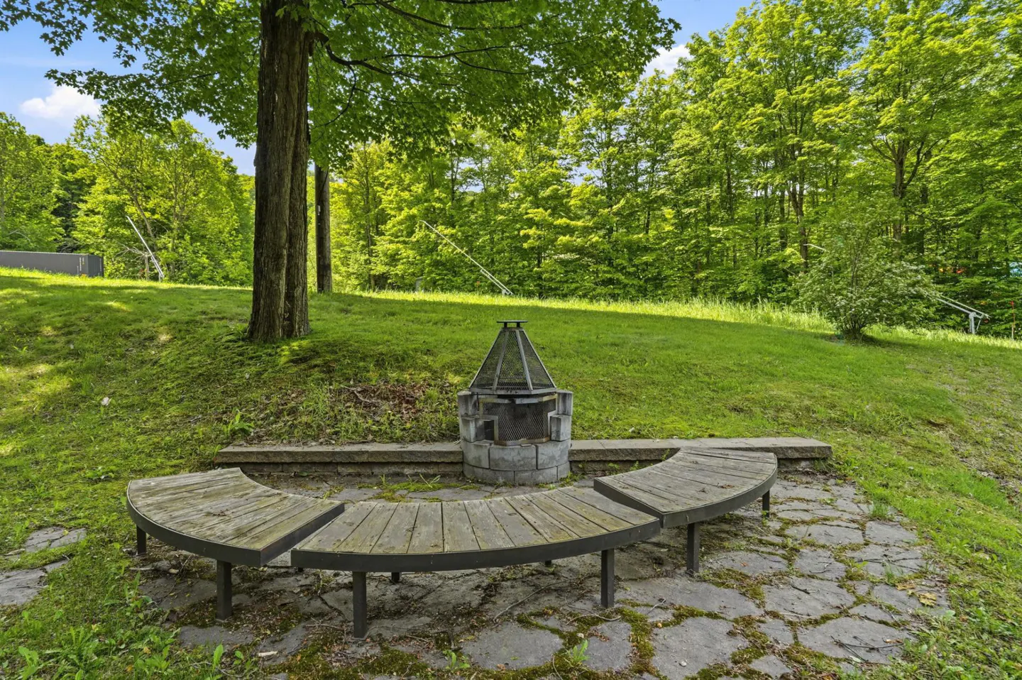 Curved wooden benches surround a stone fire pit on a flagstone patio, set against a backdrop of lush green trees.