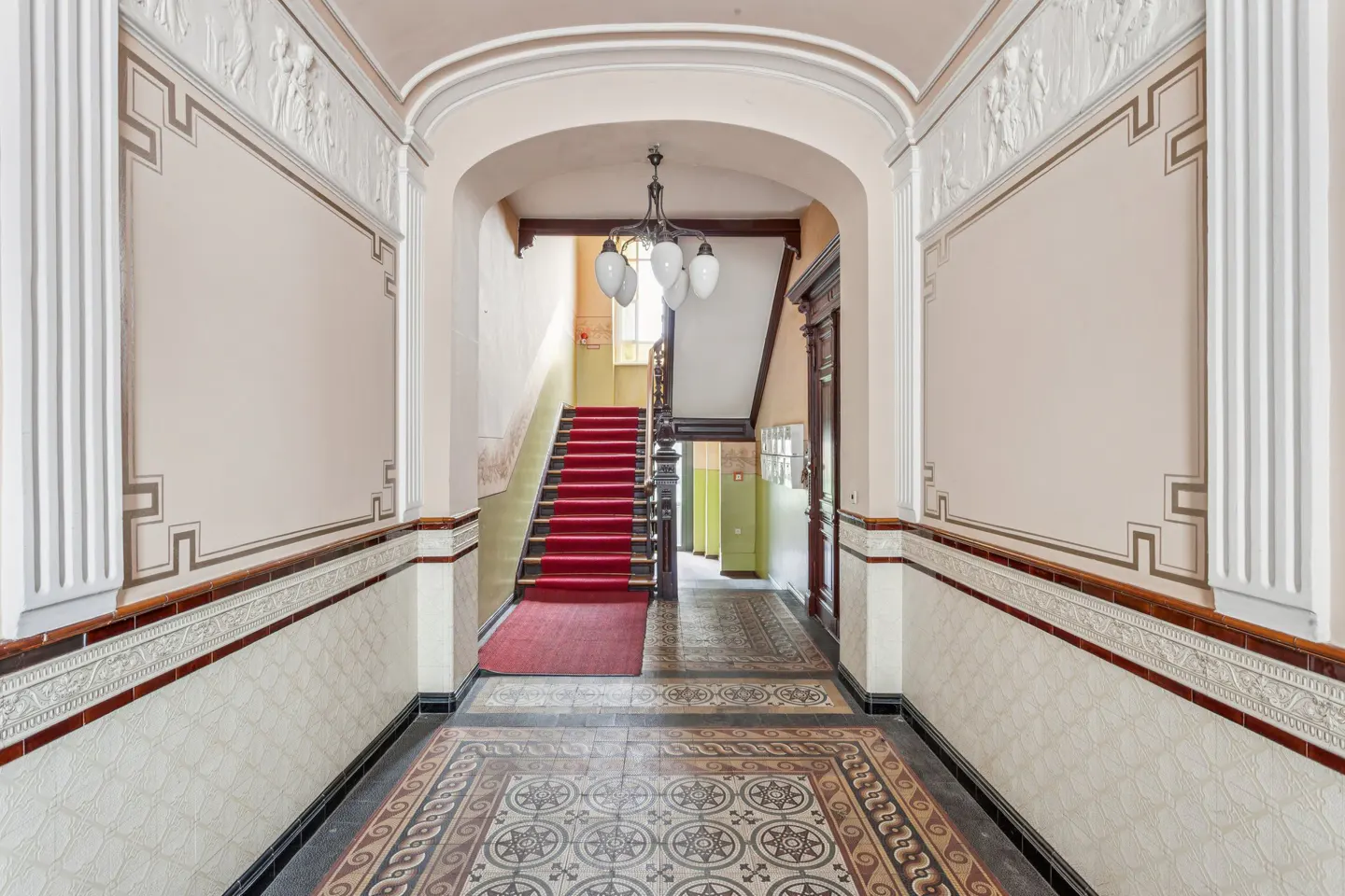 Hallway with patterned tile floor, beige walls with decorative trim, and a red-carpeted staircase leading upwards. A chandelier hangs from the arched ceiling.