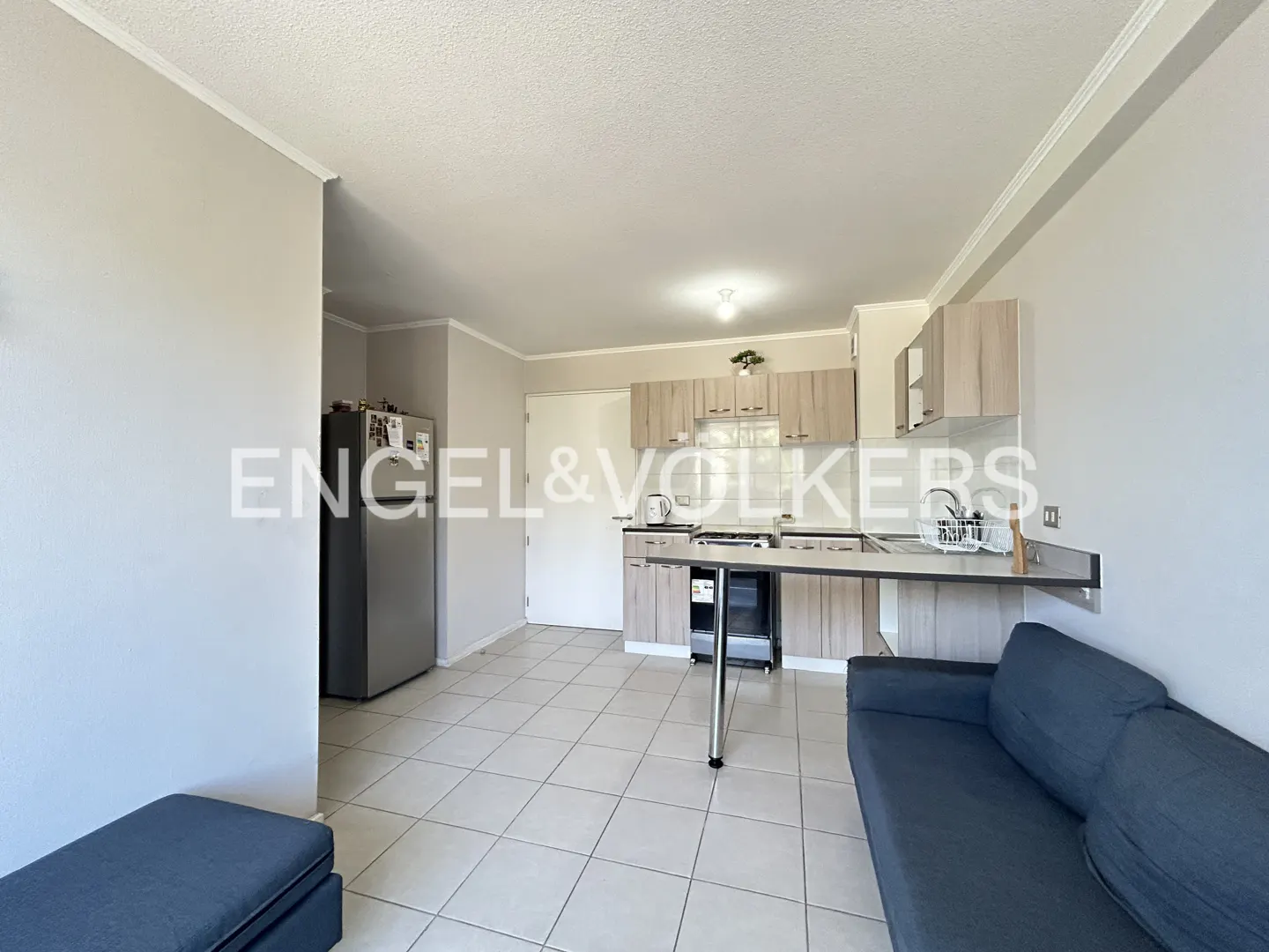 Open-plan studio apartment with a grey sofa, tiled floors, and a kitchen featuring light wood cabinets and a stainless fridge.