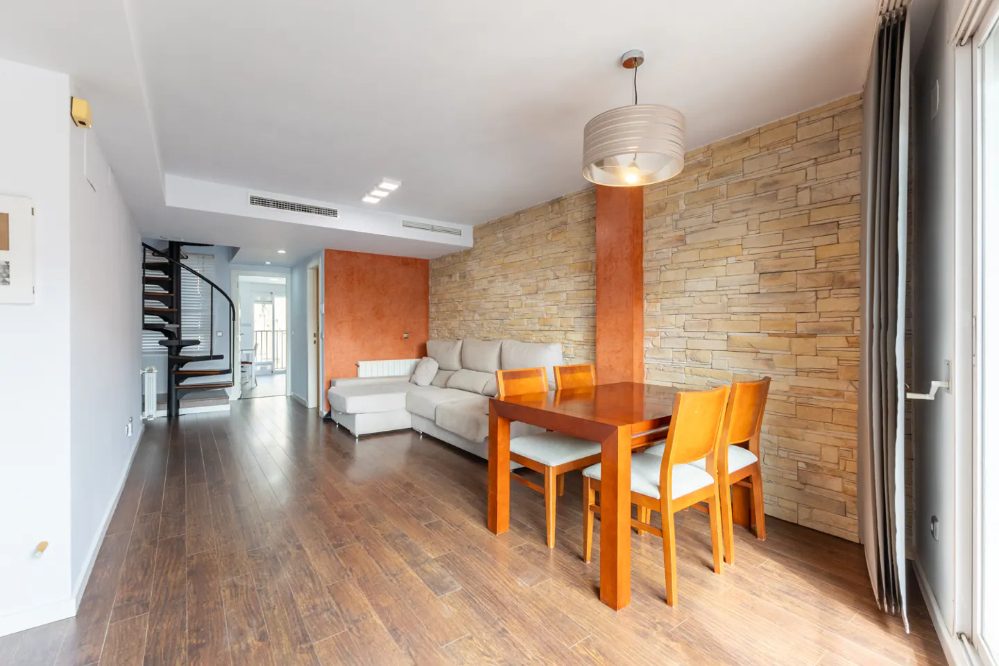 A living room with wood floors, a dining table, a beige sofa, and a spiral staircase. The walls are white, orange, and stone.