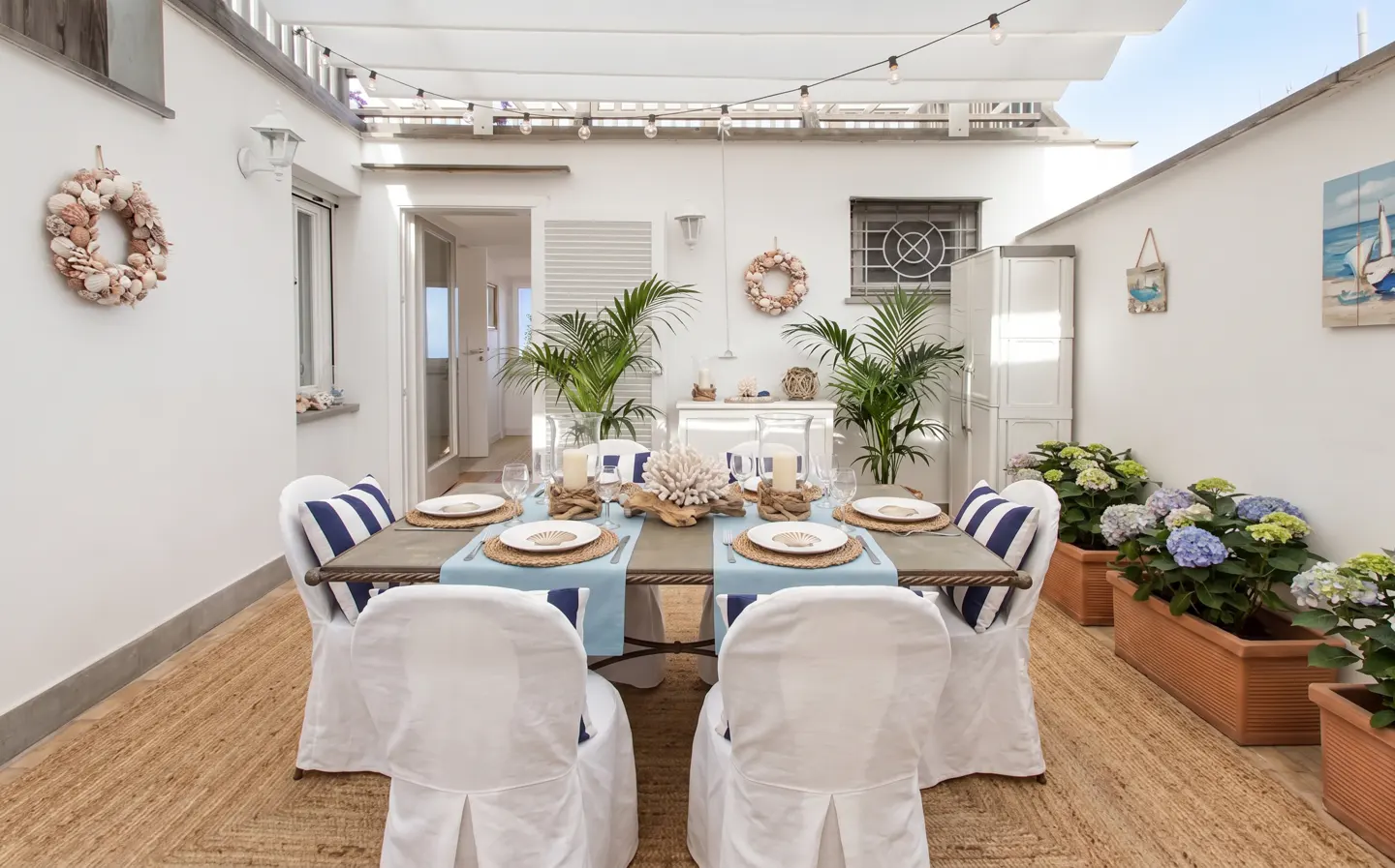 Outdoor dining area with a table set for six, white chairs, blue accents, and nautical decor. String lights hang overhead.