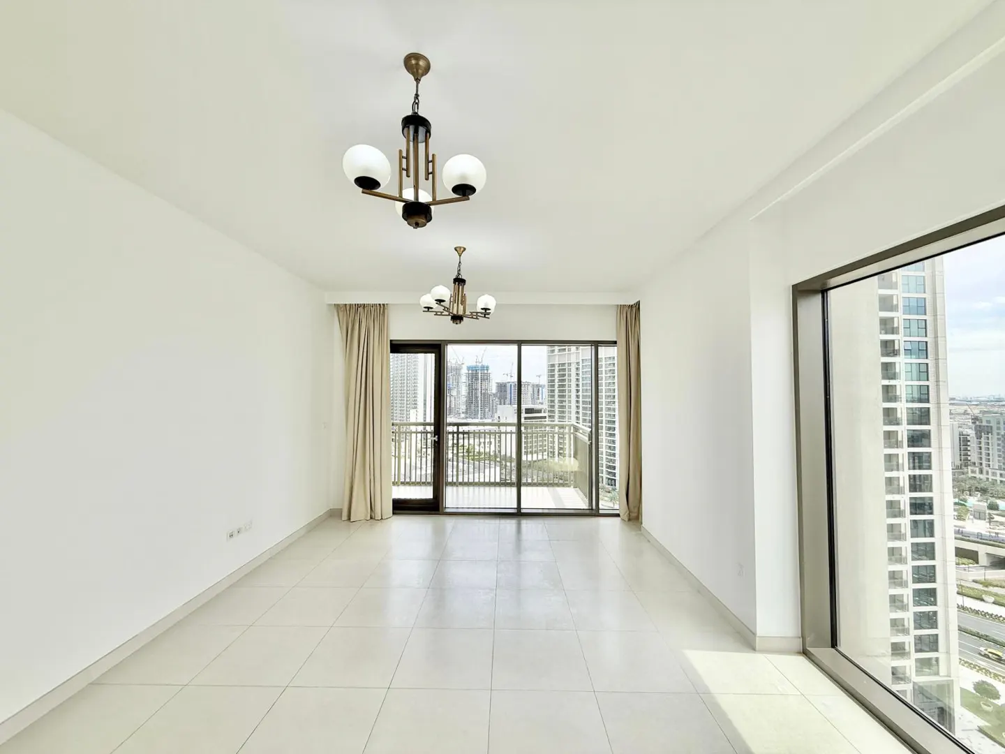 Bright, empty room with white walls and tile floor. Two chandeliers hang from the ceiling. Large windows offer a city view.