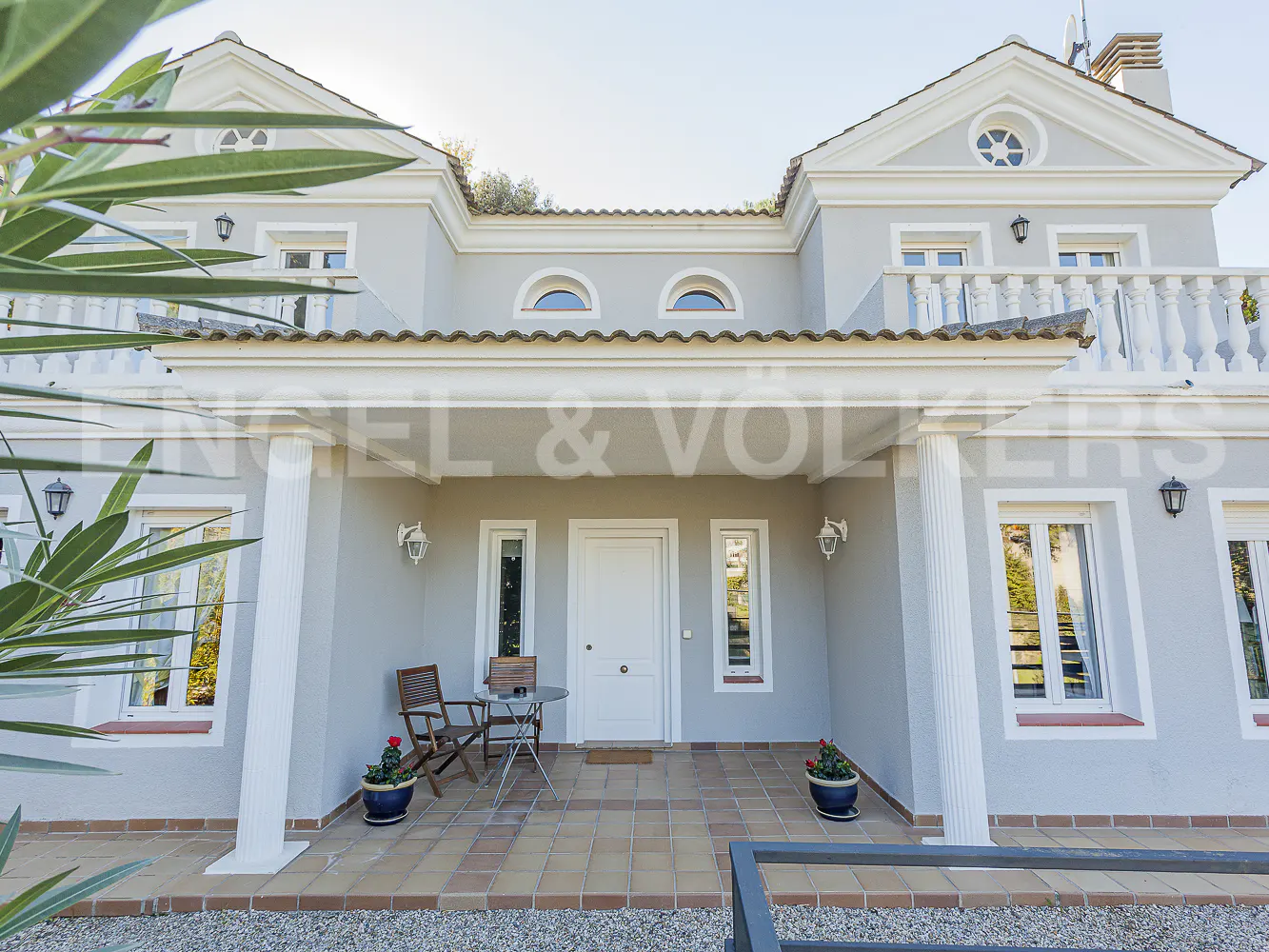Two-story gray villa with a tiled porch, white columns, a white front door, and a balcony, framed by green foliage.