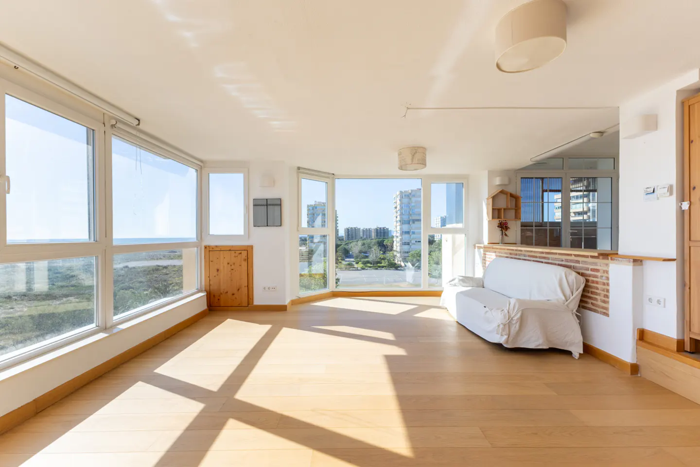 Bright, empty living room with wood floors, white walls, and large windows overlooking the ocean and city. A white sofa sits against a brick half-wall.