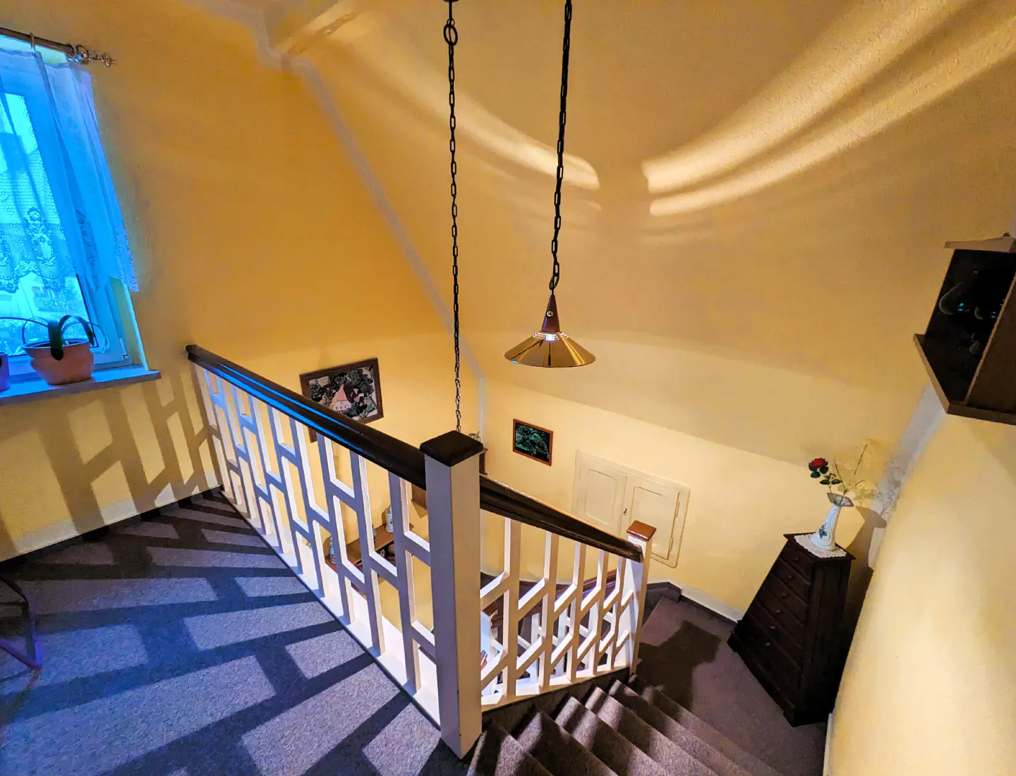 A high-angle view of a staircase with white railings and a dark handrail in a yellow room. A window with blue curtains is on the left, and a pendant light hangs from the ceiling.
